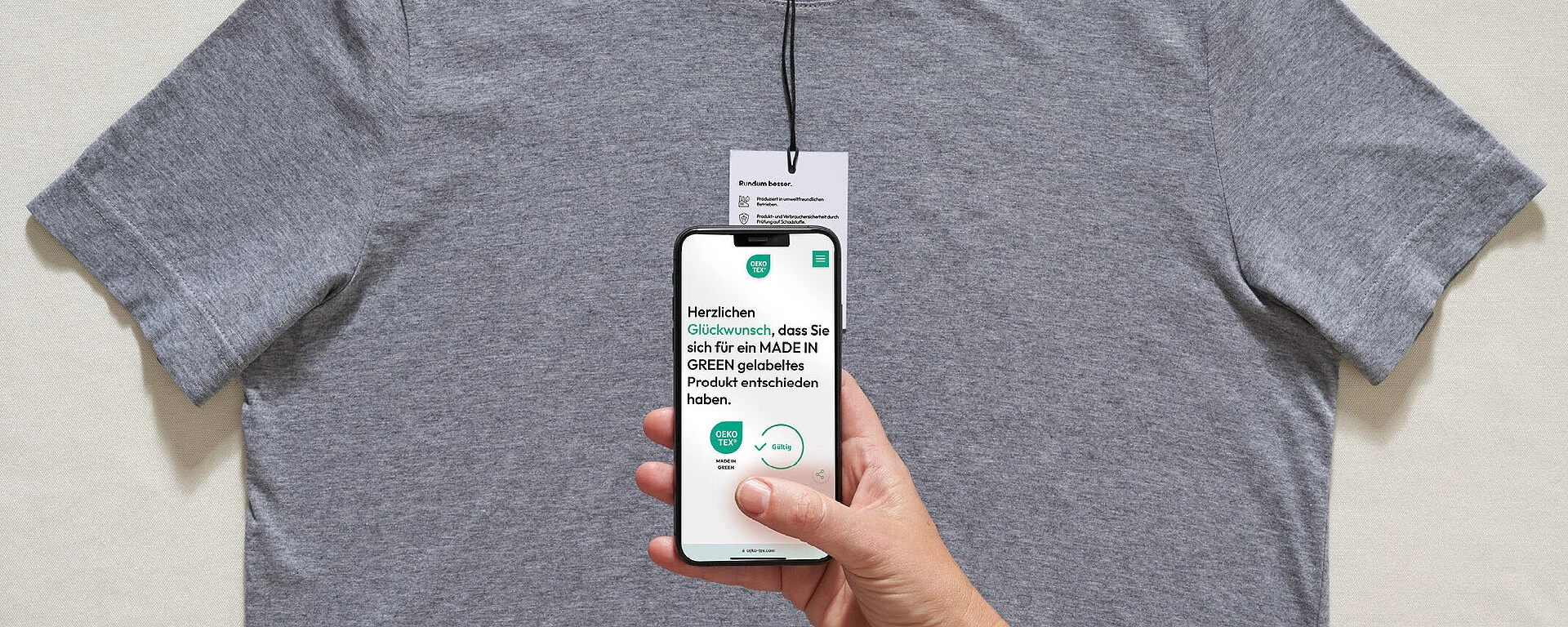 Graues T-Shirt. Hand hält Smartphone mit Glückwunsch-Nachricht für MADE IN GREEN Produktwahl