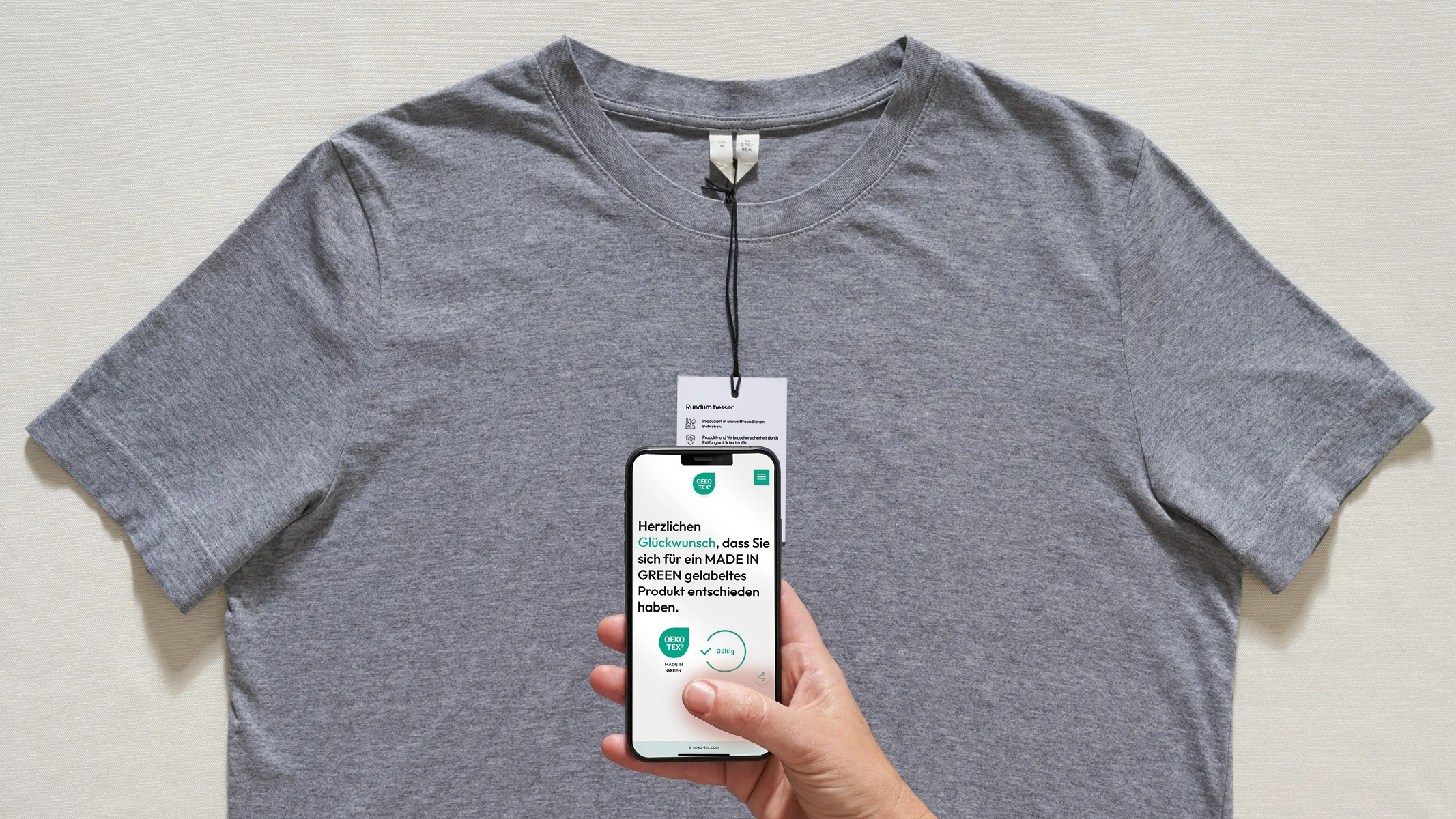 Graues T-Shirt. Hand hält Smartphone mit Glückwunsch-Nachricht für MADE IN GREEN Produktwahl
